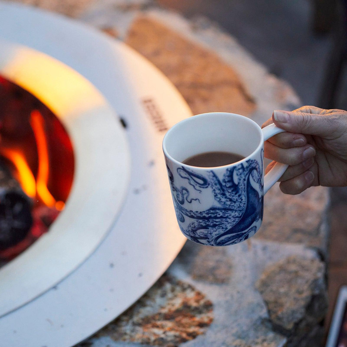 A hand holds the Lucy Mug by Caskata Artisanal Home, featuring blue designs on white porcelain, near a stone fire pit with a visible flame—perfect for enjoying your favorite coastal drinkware by the fire.