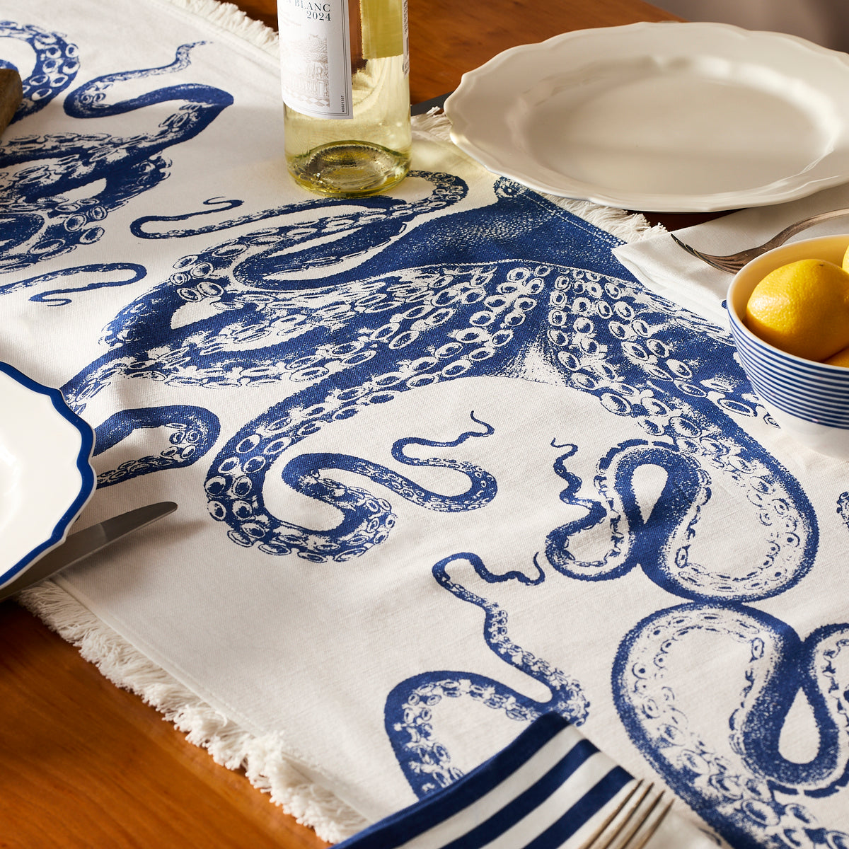 The Lucy Table Runner by Caskata adds coastal charm to a wooden table set with plates, white wine, lemons in a bowl, and striped napkins. Its blue and white octopus design brings a stylish nautical touch to your décor.