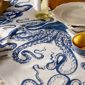 The Lucy Table Runner by Caskata, featuring a white and blue octopus print, paired with a wine bottle, plate, cutlery, and lemons creates charming coastal table decor.