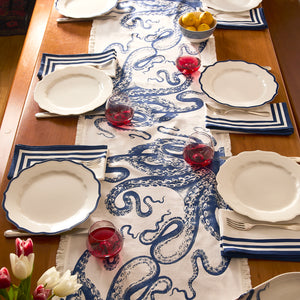 A wooden dining table with white plates, blue striped napkins, red drinks, lemons, and a tulip vase is brightened by Caskata's Lucy Table Runner—a coastal decor piece featuring a striking blue octopus motif.