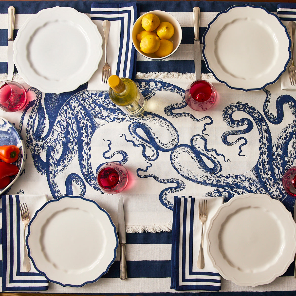 A table set for four with white plates, blue-and-white striped napkins, wine glasses, a bowl of lemons, a bottle of white wine, and Caskata’s Lucy Table Runner featuring a blue octopus for a coastal vibe.
