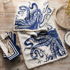 A wooden table set for coastal decor features the Caskata Lucy Table Runner, blue and white with an octopus print, styled with striped napkins, silverware, a plate, and a tray of wine glasses.
