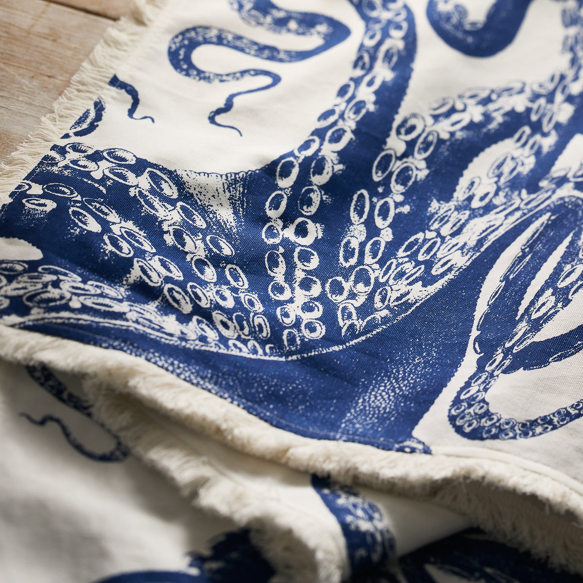 The Caskata Lucy Table Runner showcases a blue octopus tentacle print on white cotton with frayed edges—ideal for coastal table decor.