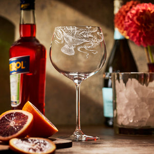 A large Caskata Lucy Gin & Tonic Glass sits empty on a bar beside a bottle of Aperol, a champagne bottle, a glass of ice, sliced blood oranges, and a vase of red flowers.