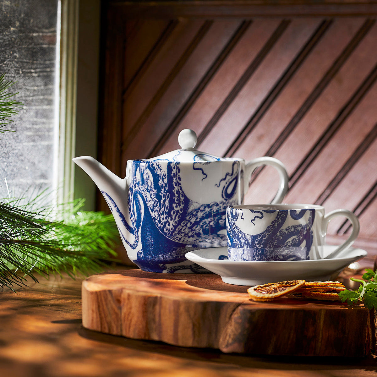 A Lucy Petite Teapot by Caskata and a hand-decorated cup sit on a wooden cutting board.