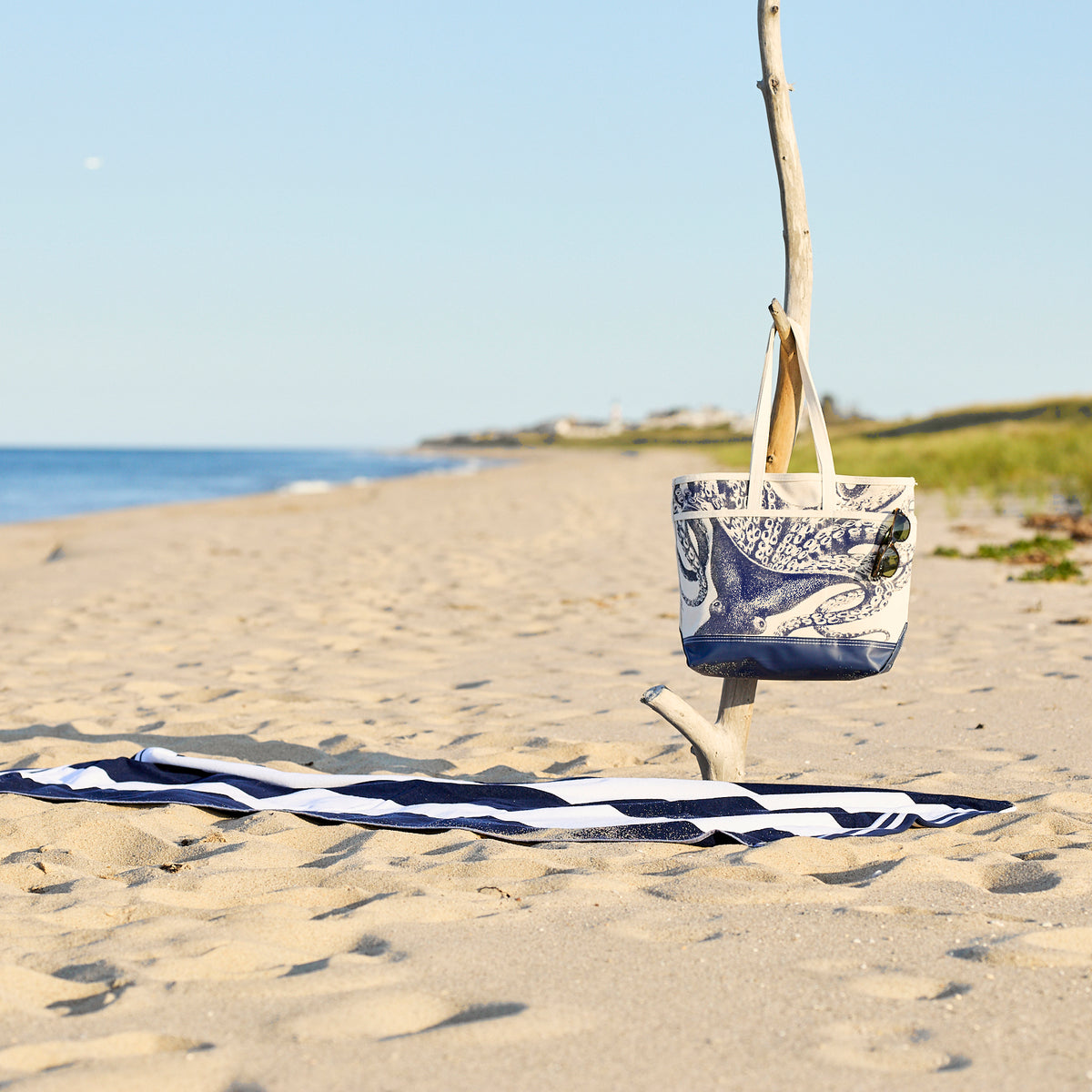 A blue and white striped towel rests on sandy beach next to a tree branch displaying the Caskata Lucy the Octopus Caskata x Steele Everyday Tote Bag, with grassy dunes and ocean in the background.