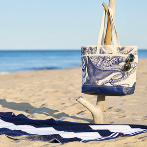 The Caskata Lucy the Octopus Caskata x Steele Everyday Tote Bag hangs on driftwood with sunglasses, a striped towel on sand beside it—all with an ocean backdrop.
