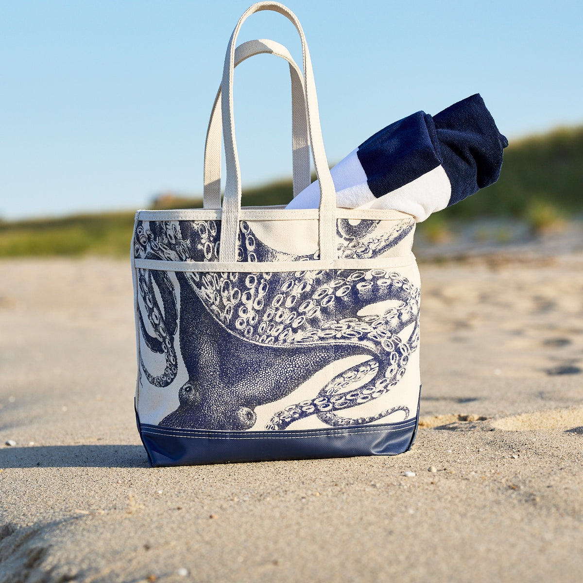 The Caskata Lucy the Octopus x Steele Everyday Tote Bag, featuring an octopus print and white straps, rests on the sand holding a rolled-up navy and white towel.