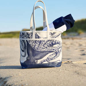 The Caskata Lucy the Octopus x Steele Everyday Tote Bag, featuring an octopus print and white straps, rests on the sand holding a rolled-up navy and white towel.
