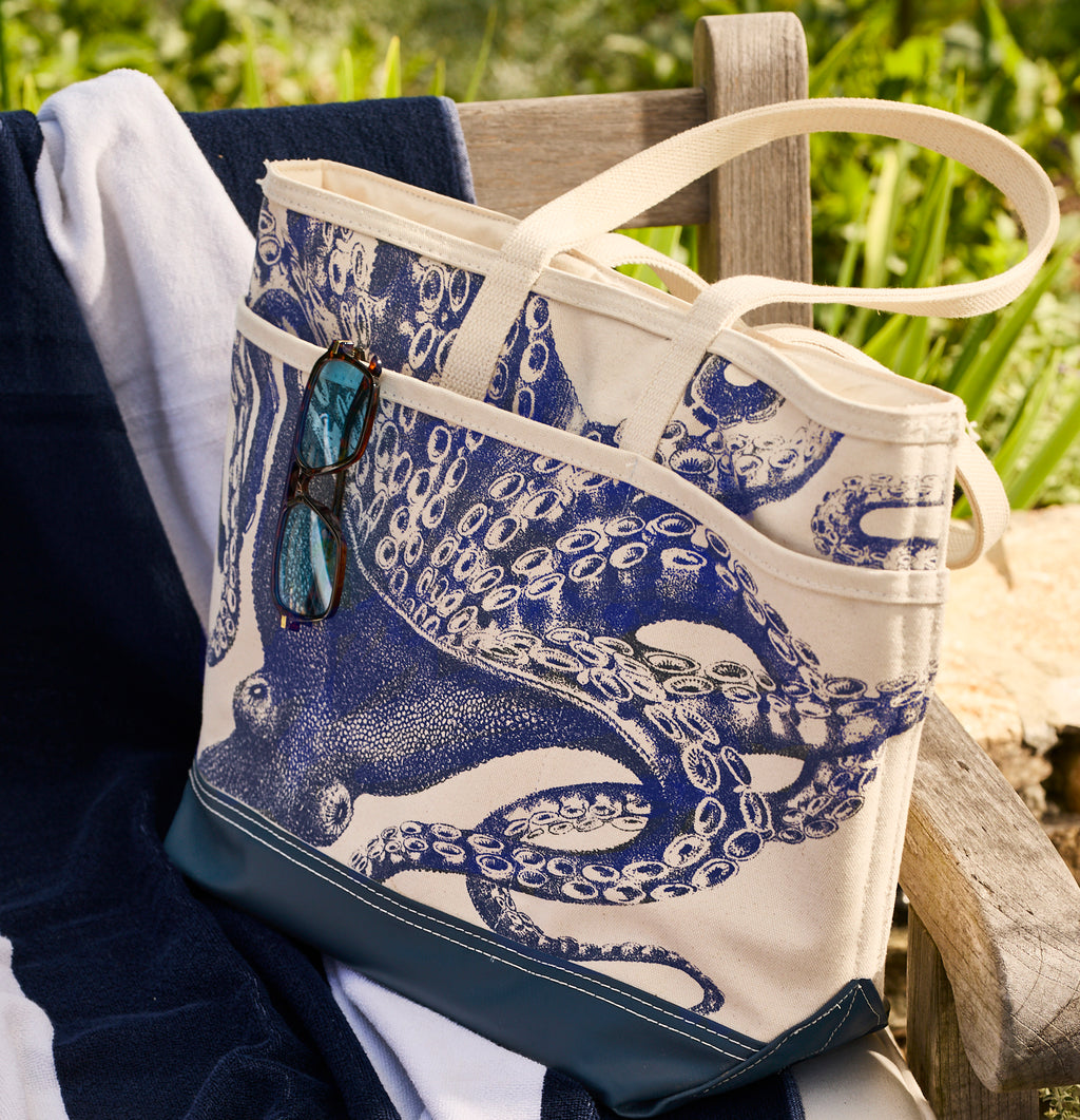 The Caskata Lucy the Octopus Caskata x Steele Everyday Tote Bag, featuring a blue octopus design and sunglasses in the pocket, sits on a striped towel atop a wooden bench outdoors.