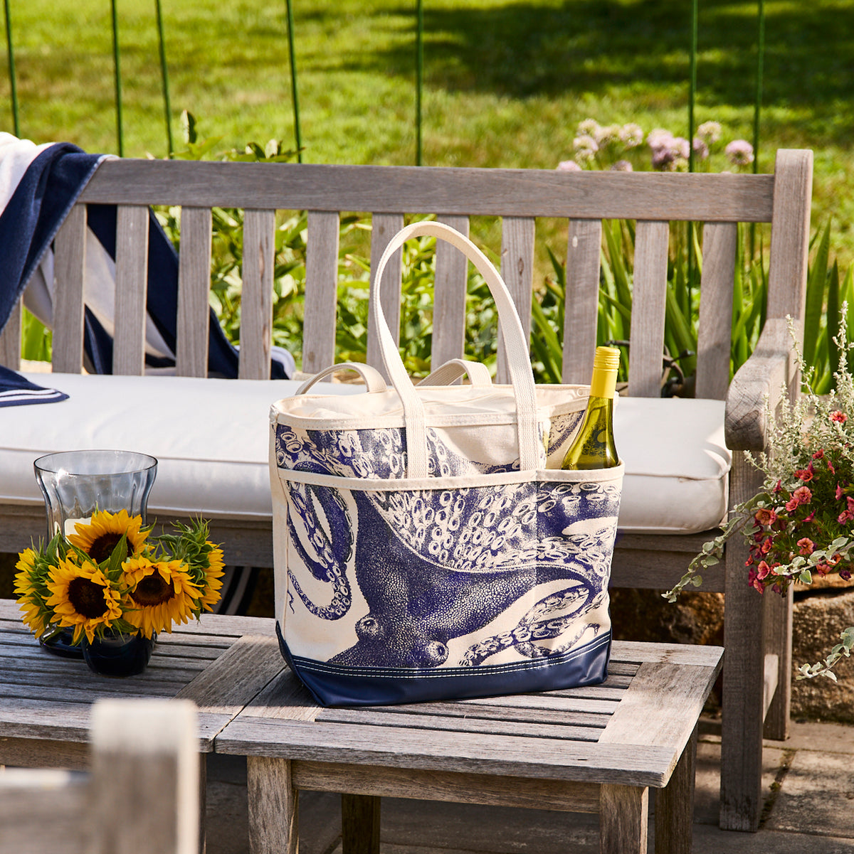 A Caskata Lucy the Octopus Caskata x Steele Everyday Tote Bag, a wine bottle, and more sit on a wooden bench. Sunflowers in a vase add cheer to the nearby table in this inviting outdoor garden scene.