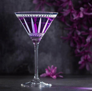 A purple cocktail in a decorative Marrakech Martini Glass from Caskata Artisanal Home, set against a dark, floral background, evokes the elegance of Art Deco with handmade crystal details.