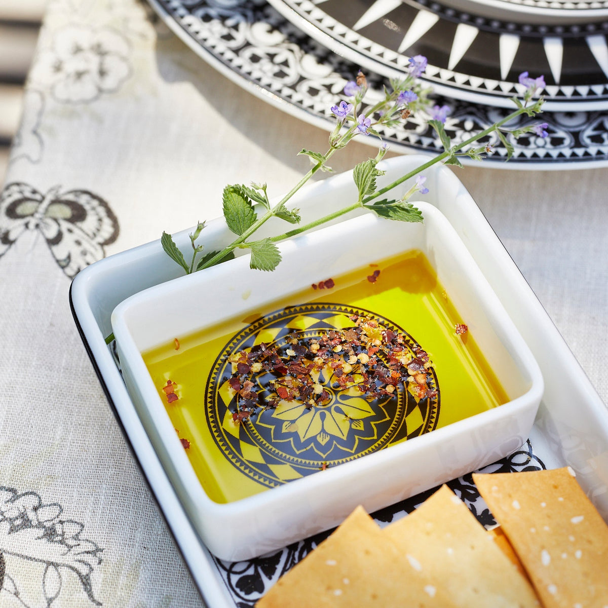 Close up of Casablanca Nested Appetizer Tray Set by Caskata. The tray is used as a dipping dish holding flavored olive oil and crackers and sits atop a floral tablecloth next to Casablanca and Marrakech dinnerware.
