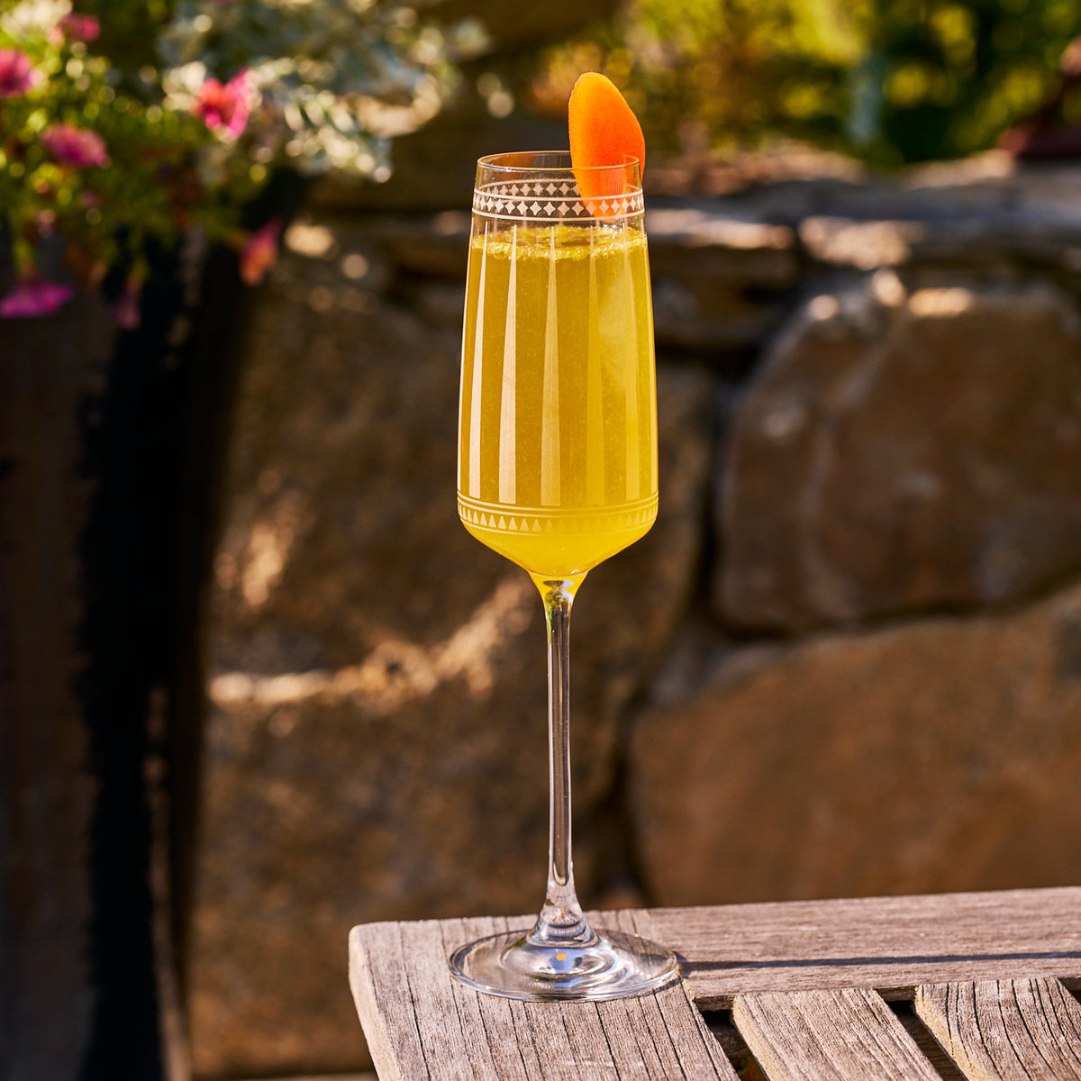 A Marrakech Champagne Glass by Caskata Artisanal Home, filled with a yellow-orange cocktail and garnished with an orange slice, rests on a wooden outdoor table against a stone wall backdrop.