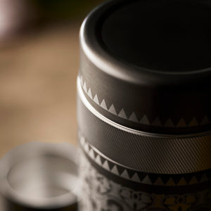 A close-up of Caskata’s The Golden Hour Bar Bundle shows a cylindrical metal cocktail shaker with intricate patterns and a zigzag band near the top; another matching piece of barware is softly blurred in the background.
