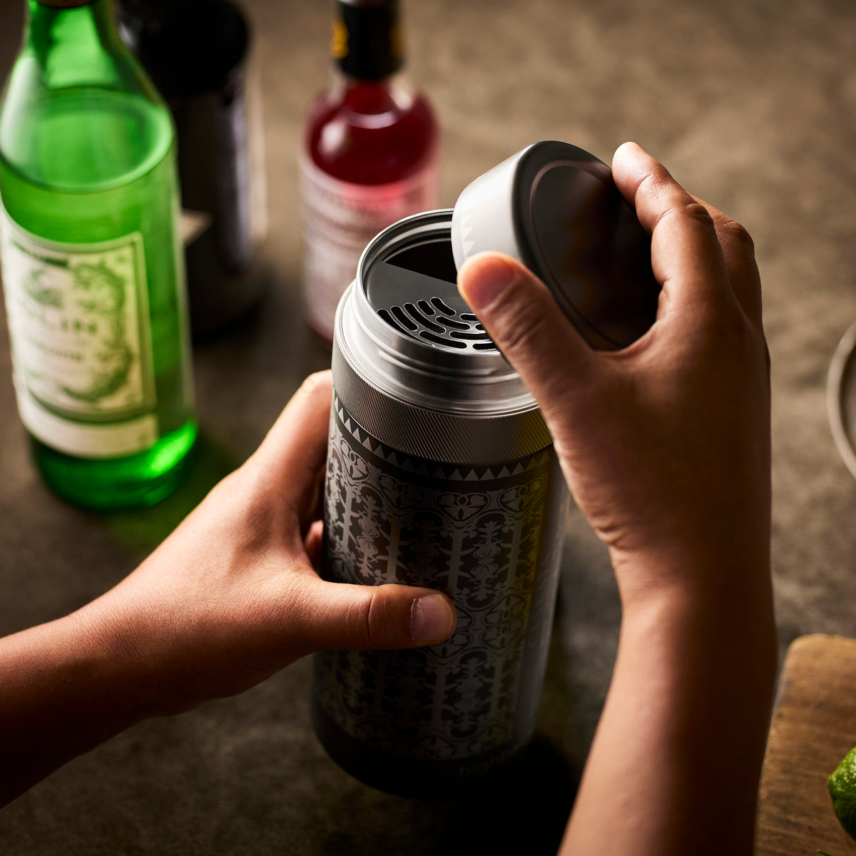 Someone opens the lid of Caskata’s The Art Deco Bar Bundle cocktail shaker, with bottles of spirits, mixers, and stemless wine glasses displayed on a countertop in the background.