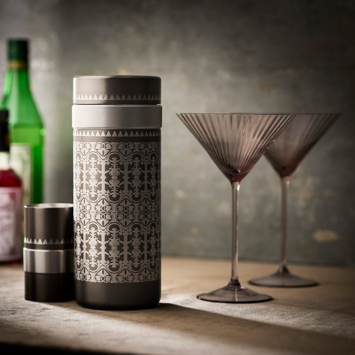 A patterned Caskata Casablanca Cocktail Shaker and lid rest beside two empty martini glasses, perfect for making Quinn Mocha Martinis, while bottles in the background suggest the complete Smoky Quartz Bar Bundle.