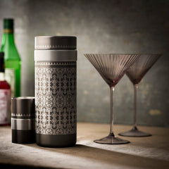 A patterned Caskata Casablanca Cocktail Shaker and lid rest beside two empty martini glasses, perfect for making Quinn Mocha Martinis, while bottles in the background suggest the complete Smoky Quartz Bar Bundle.