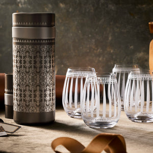 The Art Deco Bar Bundle by Caskata features a metallic canister with intricate patterns paired with four etched stemless wine glasses, displayed on a neutral-toned surface.