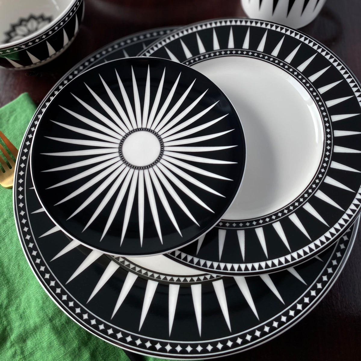 Stacked Marrakech Small Plates with a black and white geometric starburst design on a dark surface, accompanied by a green cloth and a gold fork. These high-fired porcelain pieces from Caskata Artisanal Home evoke the charm of Marrakech.