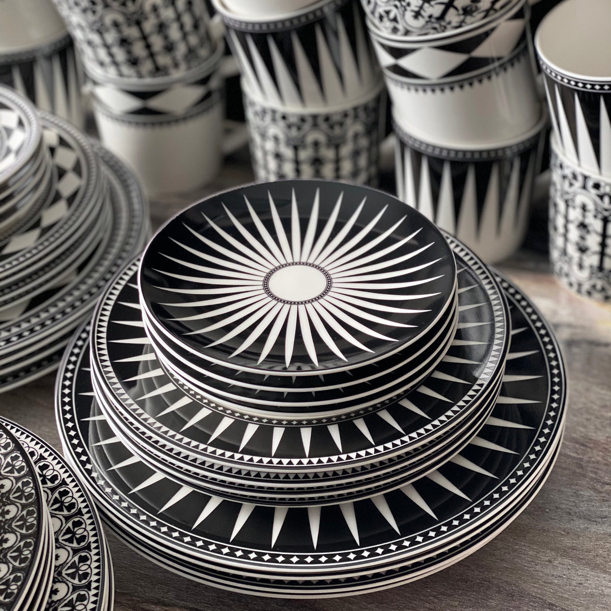 A stack of black and white patterned Marrakech Small Plates by Caskata Artisanal Home and high-fired porcelain bowls arranged on a wooden surface, with similarly patterned cups in the background evokes the charm of Marrakech.