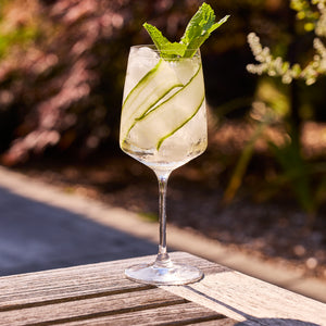 A refreshing cocktail with ice, cucumber, and mint in a Caskata Artisanal Home Marrakech White Wine Glass sits on an outdoor wooden table, garden softly blurred in the background.