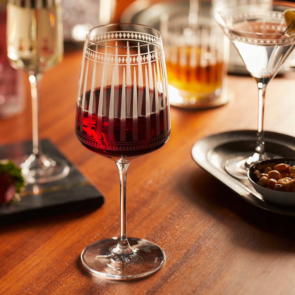 The "Cocktail Hour" Bundle by Caskata includes a glass of red wine on a wooden table next to an appetizer tray, with mixed nuts and other drinks in the background.