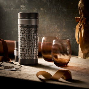 The Golden Hour Bar Bundle by Caskata, featuring a patterned metal flask and two ribbed brown glasses, rests on a textured surface with a ribbon, gift tag, and wrapped present nearby.