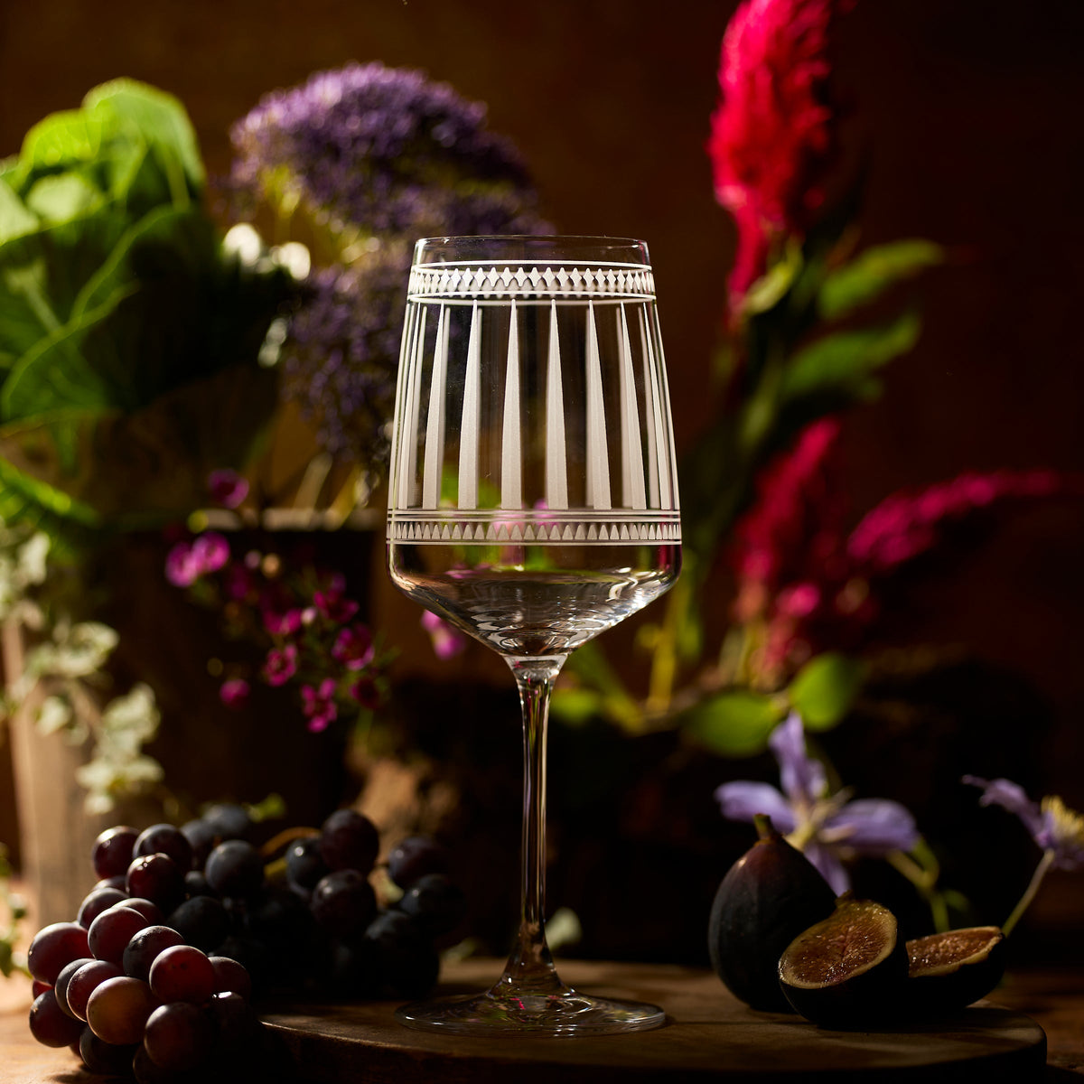 A Marrakech Red Wine Glass by Caskata Artisanal Home, crafted from clear lead-free crystal with a decorative rim, stands on a table with grapes, figs, and flowers in the background.