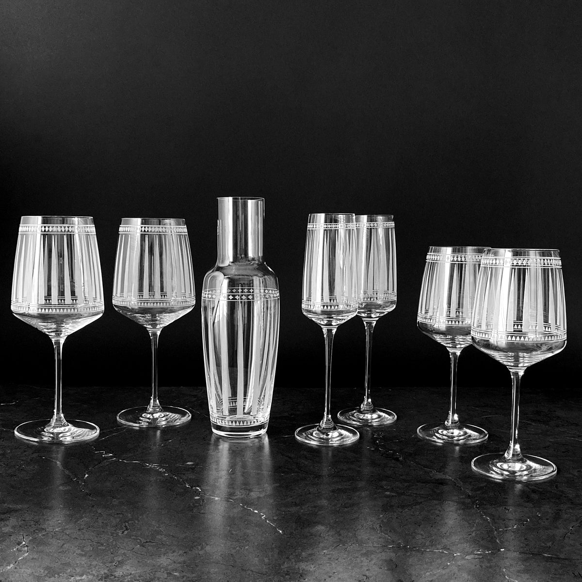 A black and white photo of a set of Marrakech Carafe wine glasses and a Caskata Artisanal Home decanter with graphic patterns inspired by Art Deco, reminiscent of the stylish elegance found in Marrakech.