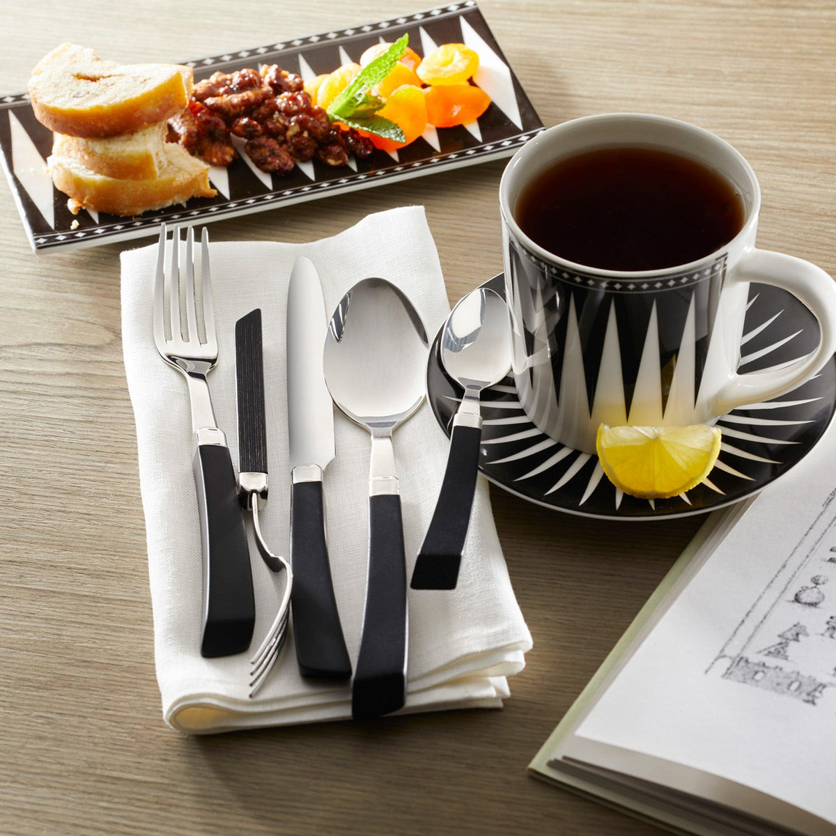 On a wooden table, a cup of tea in exquisite bone china with a lemon slice rests on a saucer beside cutlery and napkin. Slices of bread and nuts with fruit are elegantly arranged on the Marrakech Petite Serving Tray by Caskata, completing this Art Deco-inspired setting.