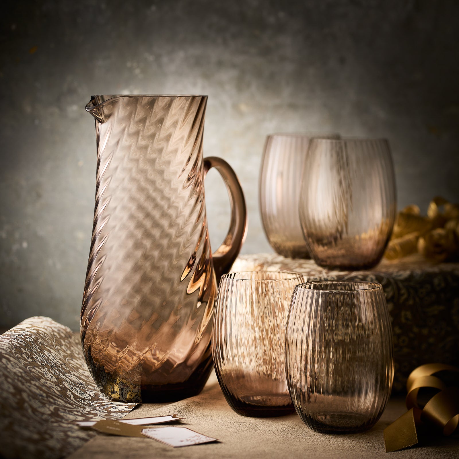 The Caskata "Moody Mocha" Bundle features a handblown glass pitcher and four matching muted pink tumblers—an elegant gift set, beautifully arranged with decorative paper and ribbon for any special occasion.