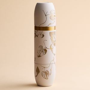 The Caskata x High Camp Crema Morning Glory Fern Wine Flask by Caskata, featuring a white cylindrical form adorned with a gold band and metallic leaf pattern, stands upright against a beige background, exuding refined elegance reminiscent of its stainless steel build paired gracefully with magnetized tumblers.