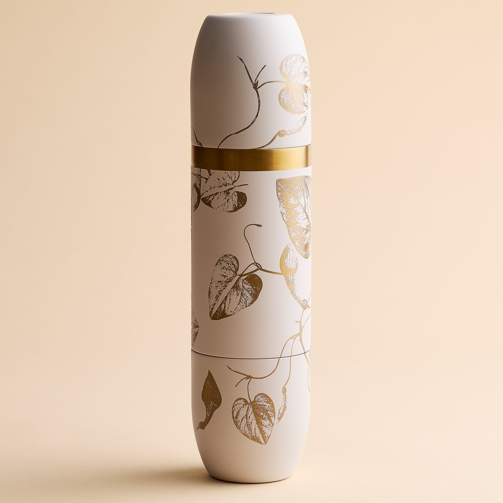 The Caskata x High Camp Crema Morning Glory Fern Wine Flask by Caskata, featuring a white cylindrical form adorned with a gold band and metallic leaf pattern, stands upright against a beige background, exuding refined elegance reminiscent of its stainless steel build paired gracefully with magnetized tumblers.