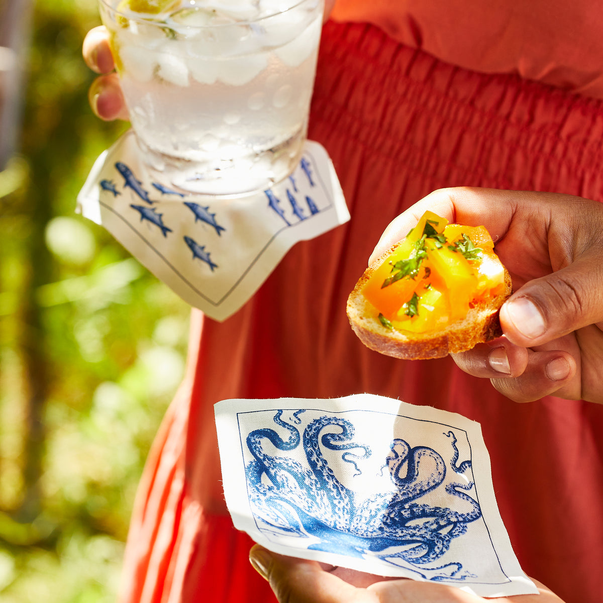 A person in an orange dress enjoys a drink and bruschetta outdoors with Caskata’s “Ella in the Morning” Bundle—featuring Spiky Fish Mug, fish-patterned napkin, and octopus Coastal Cocktail Napkin Roll—perfect for any beach lover.
