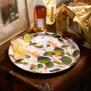 The Caskata Nasturtiums Large Round Birchwood Tray, handmade in Finland, holds a glass of ice and lime, a bottle of liquor, sunglasses, and a brown paper bag on a leather-topped chest.