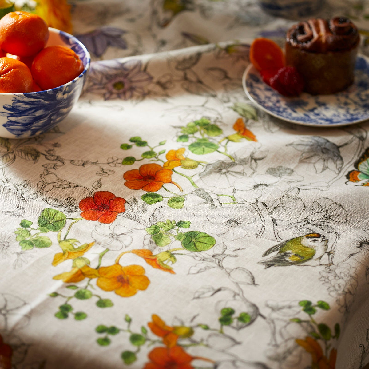 A Nasturtium Linen Tablecloth from TTT adorned with beautiful flowers and fresh fruit.