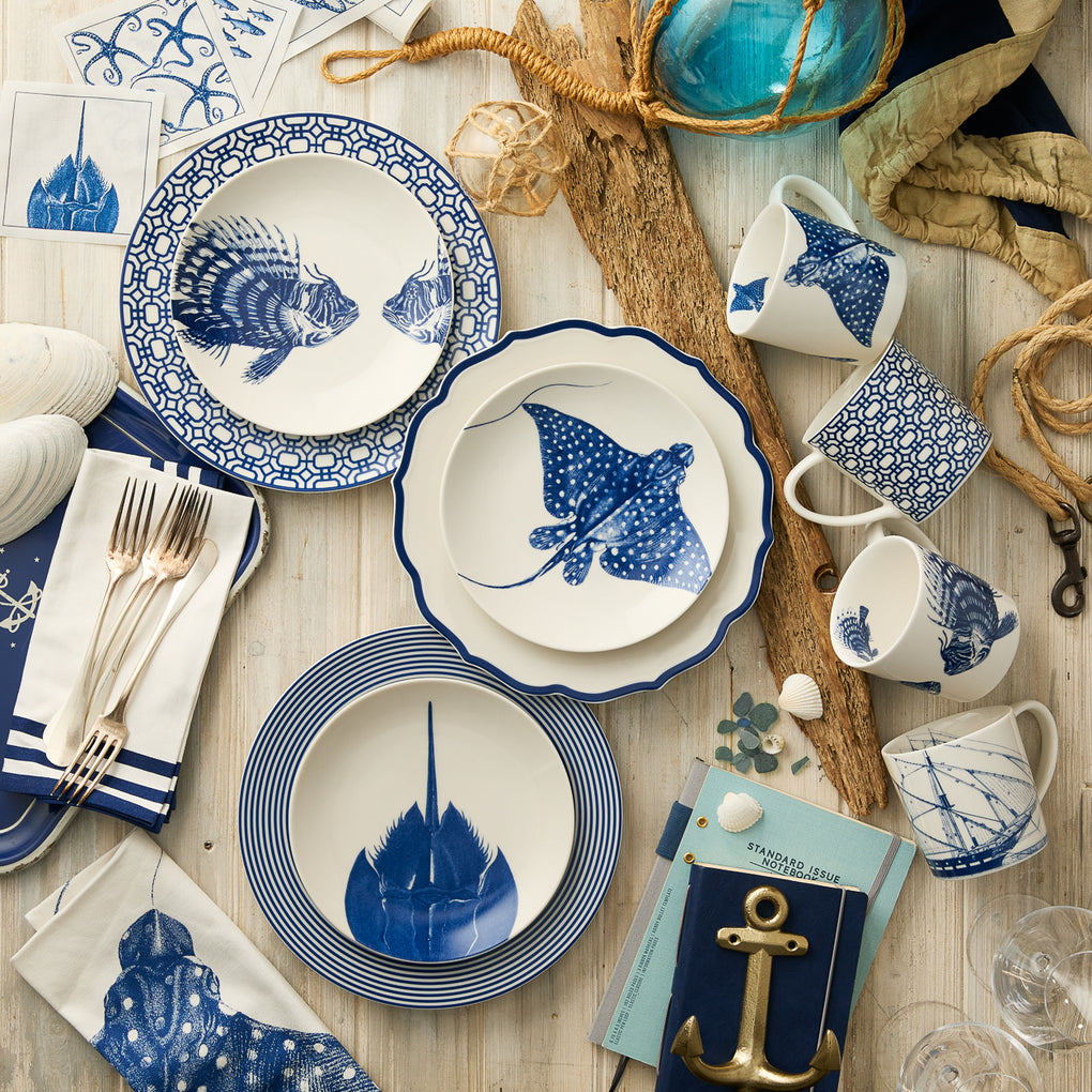 A nautical table setting with Caskata Artisanal Home Horseshoe Crab Coupe Salad Plates, blue and white dinnerware, mugs, bowls with marine motifs, silverware, napkins, seashells, a rope accent, driftwood pieces, and gold anchor decor.