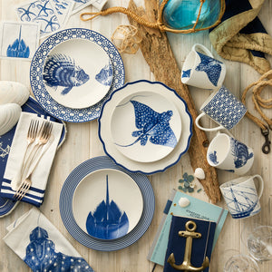 A nautical table setting with Caskata Artisanal Home Horseshoe Crab Coupe Salad Plates, blue and white dinnerware, mugs, bowls with marine motifs, silverware, napkins, seashells, a rope accent, driftwood pieces, and gold anchor decor.