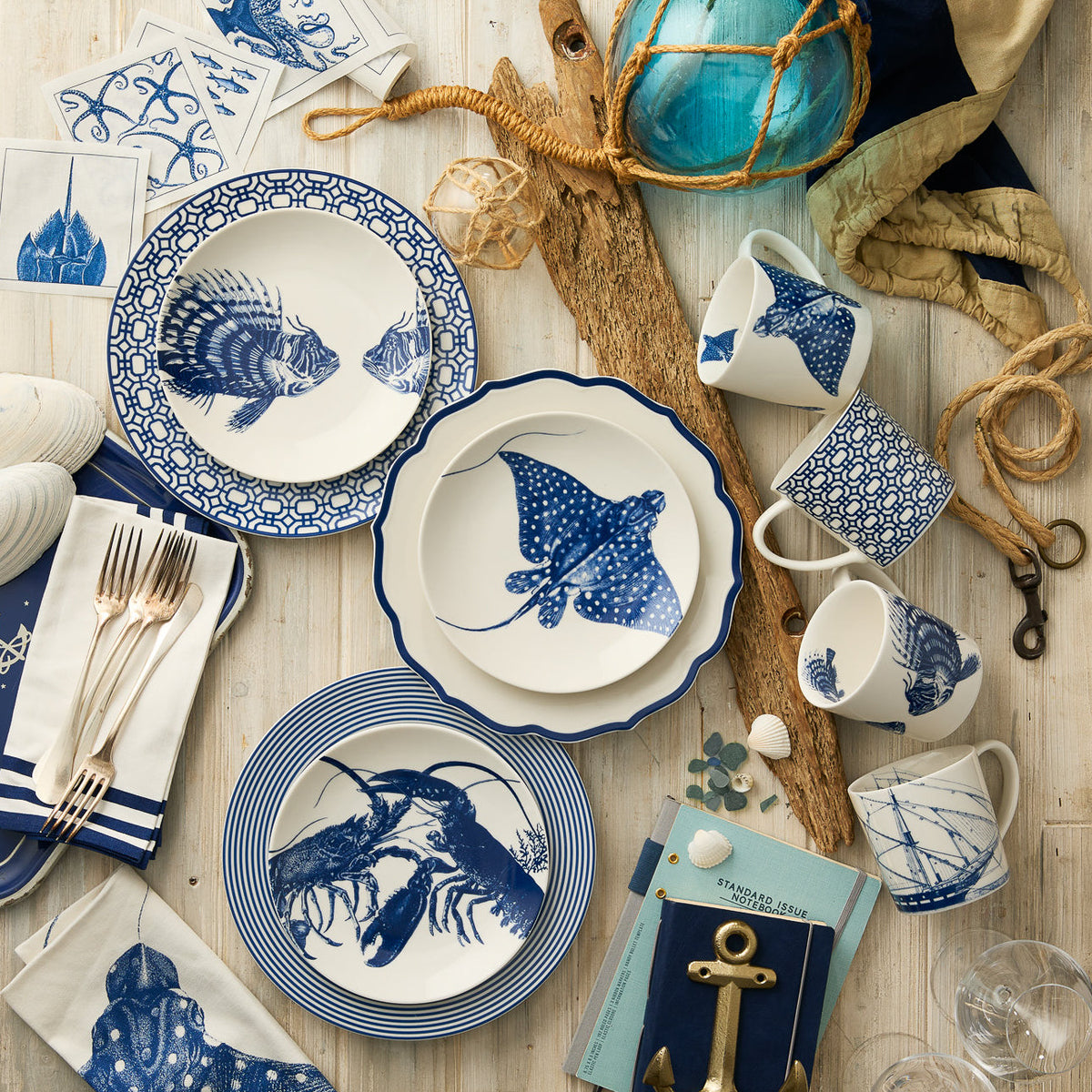 Create a coastal table setting with Caskata's Lobster Coupe Salad Plate, featuring sea life designs in blue and white, complemented by nautical decor like rope, an anchor, and shells.