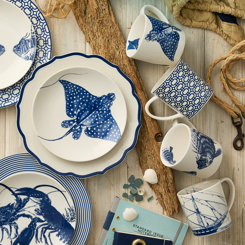 Caskata's "Ray" the Stingray Mug is displayed on a light wooden surface among assorted blue and white ceramic plates, coastal accents, nautical props, and seashells.