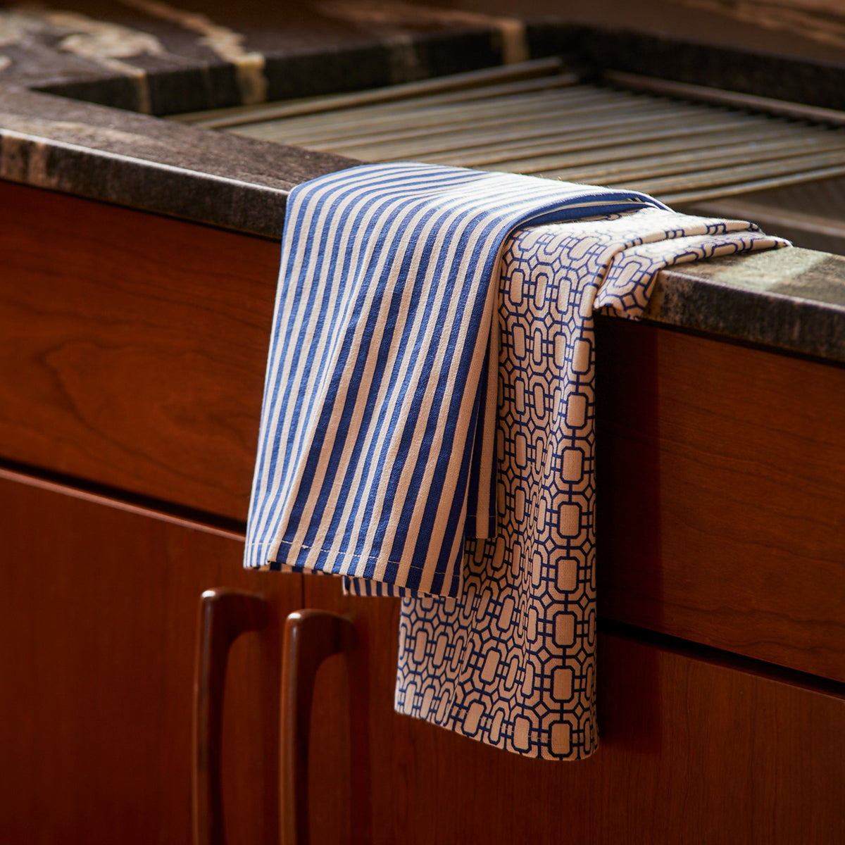 Two Newport Garden Gate &amp; Pinstripe Blue Kitchen Towels, Mixed Set of 2 by Caskata, hang over the edge of a dark countertop. Brown wooden cabinets are below.