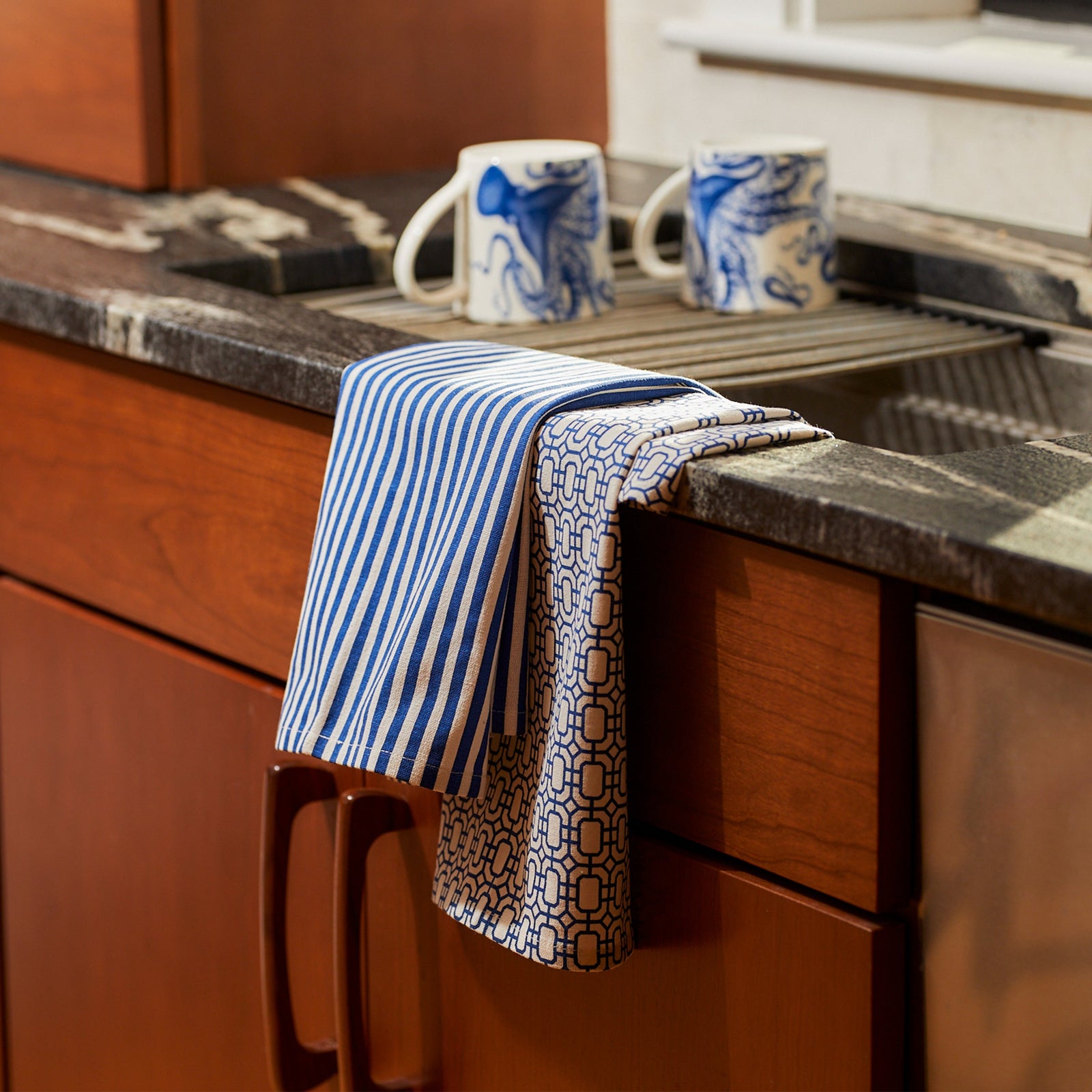 Two folded kitchen tea towels: one with blue and white stripes, and the other with a Newport Garden Gate-inspired blue geometric chain pattern on a white background. Both are made from 100% cotton, ensuring durability and style. These are the Newport Garden Gate & Pinstripe Blue Kitchen Towels, Mixed Set of 2 by Caskata.