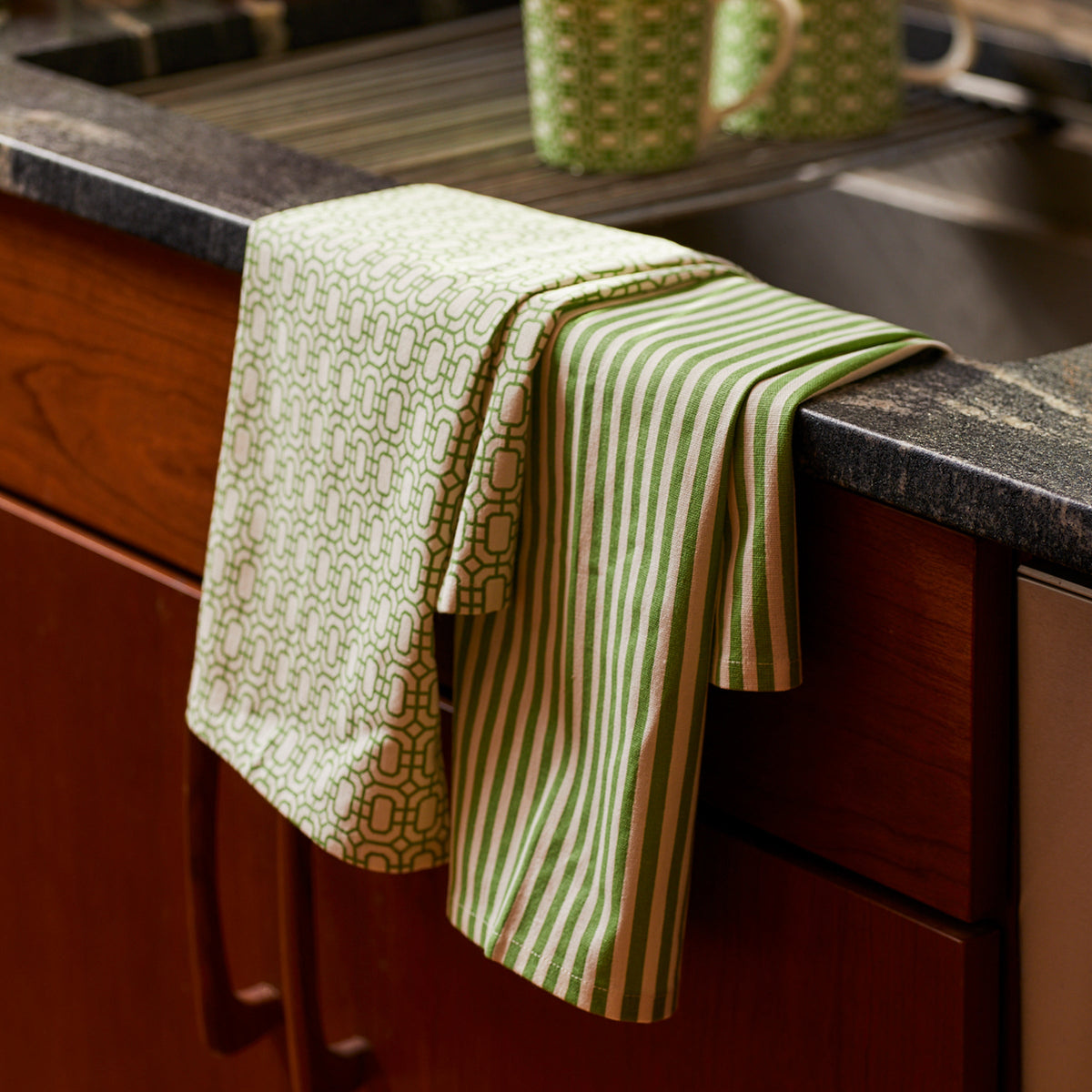 A green and white patterned kitchen towel, part of our Newport Garden Gate &amp; Pinstripe Verde Kitchen Towels, Mixed Set of 2 collection by Caskata, is draped over the edge of a sink with wooden cabinets below. Nearby, a ceramic mug with a similar Newport Garden Gate pattern sits on the counter, making perfect kitchen companions.