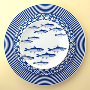 Three nested plates featuring different blue and white patterns; the top plate depicts a school of fish, the middle plate a geometric pattern, and the bottom plate has concentric stripes. The School of Fish Small Plates from Caskata Artisanal Home are microwave safe, heirloom-quality dinnerware that brings both beauty and practicality to any table setting.