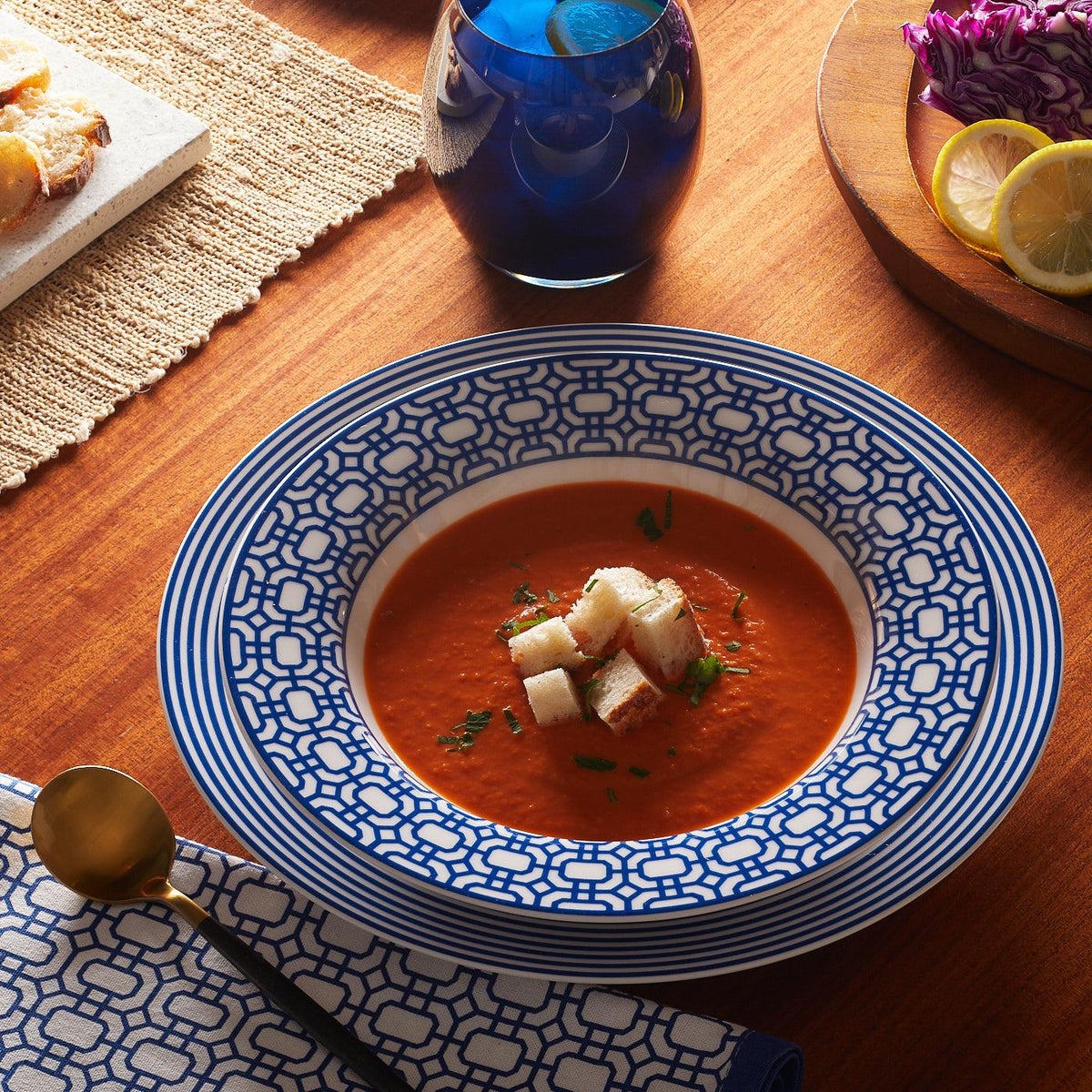 A bowl of tomato soup garnished with croutons and herbs is placed on a wooden table, accompanied by contemporary dinnerware including a Caskata Artisanal Home Newport Stripe Rimmed Dinner Plate, a gold spoon, napkin, bread, purple cabbage, and lemon slices.