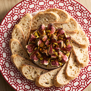The Newport Crimson Garden Gate Table for 4 by Caskata is set for entertaining, with a wheel of cheese topped with figs and pistachios surrounded by baguette slices, perfect to complement your dinnerware.