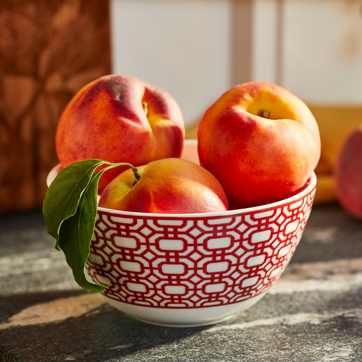 Four ripe peaches—one with a green leaf—rest in a white bowl with a red geometric pattern atop the sunlit Caskata Newport Crimson Garden Gate Table for 4.