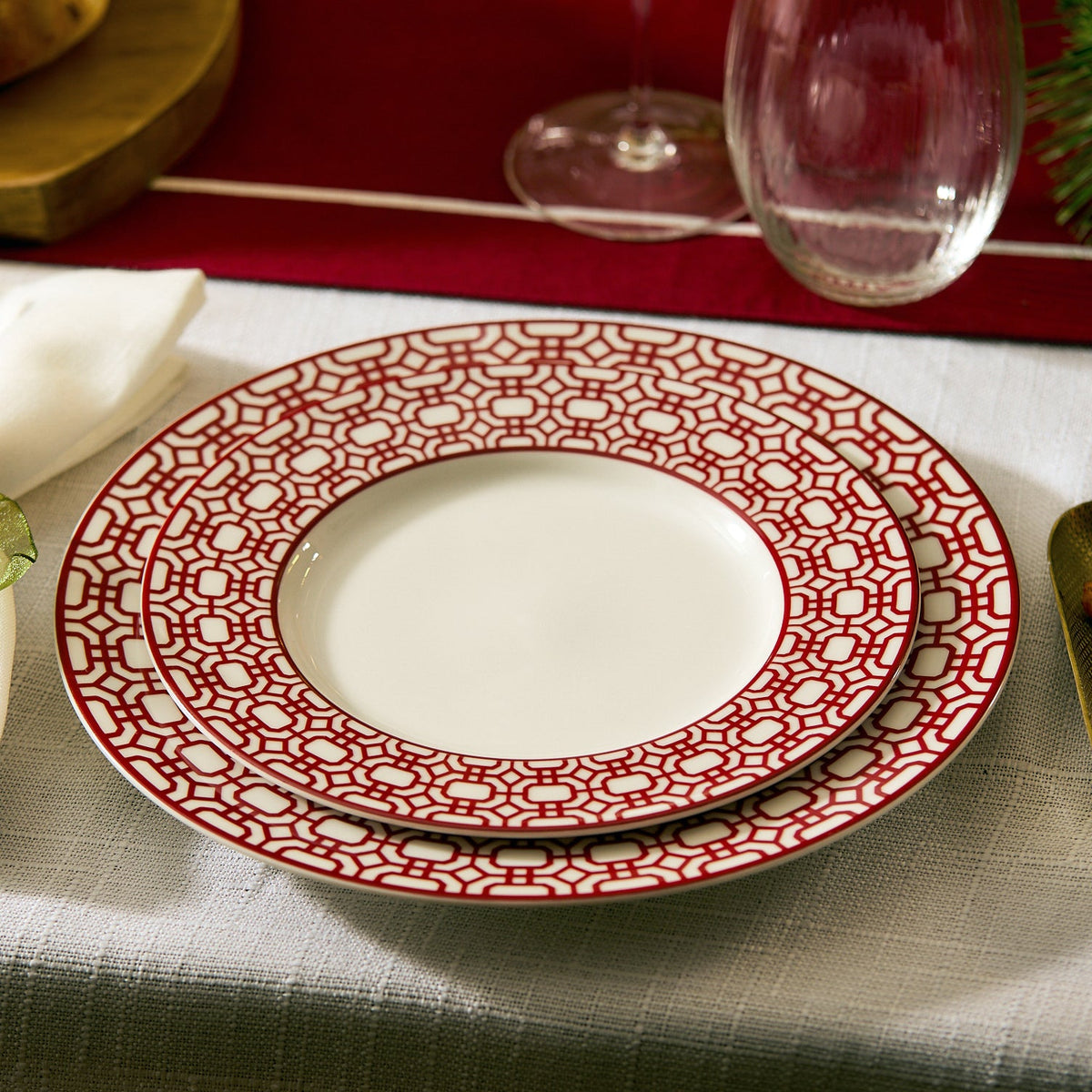Two white plates from Caskata’s Newport Crimson Garden Gate Table for 4, with a red geometric design, are stacked on a white tablecloth next to a wine glass and napkin—a stylish porcelain addition to your dining table.