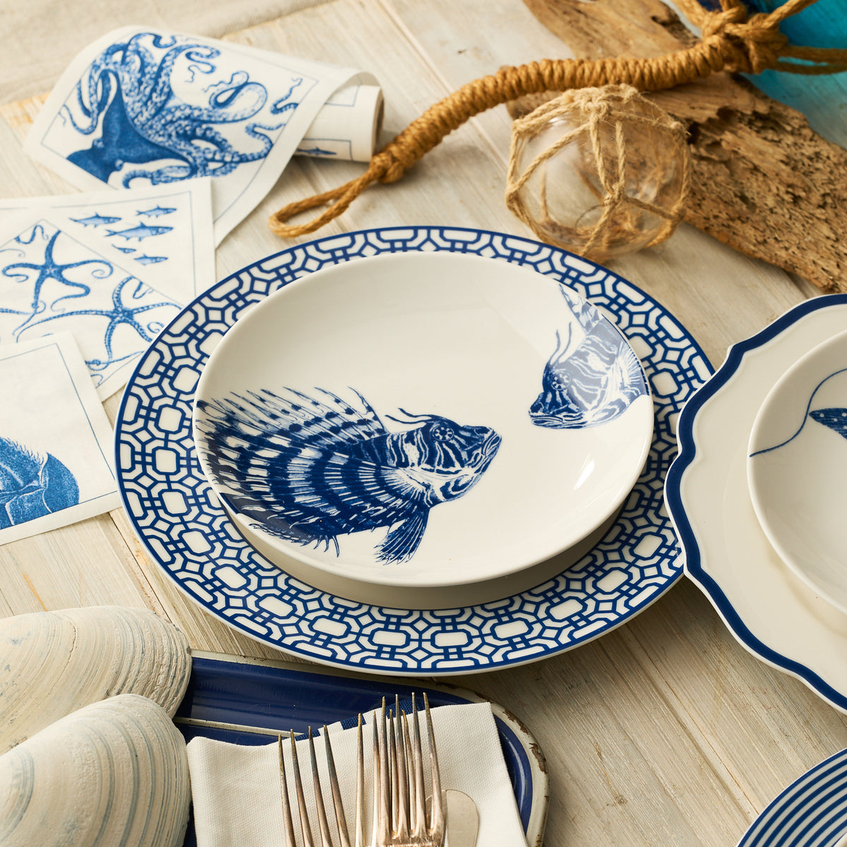 The Caskata Spiky Fish Coupe Salad Plate, featuring a blue fish design, rests on a patterned dinner plate on a wooden table set with nautical-themed decor and cutlery.